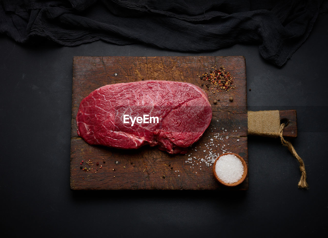 Raw beef tenderloin lies on a cutting board and spices for cooking on a black table, top view