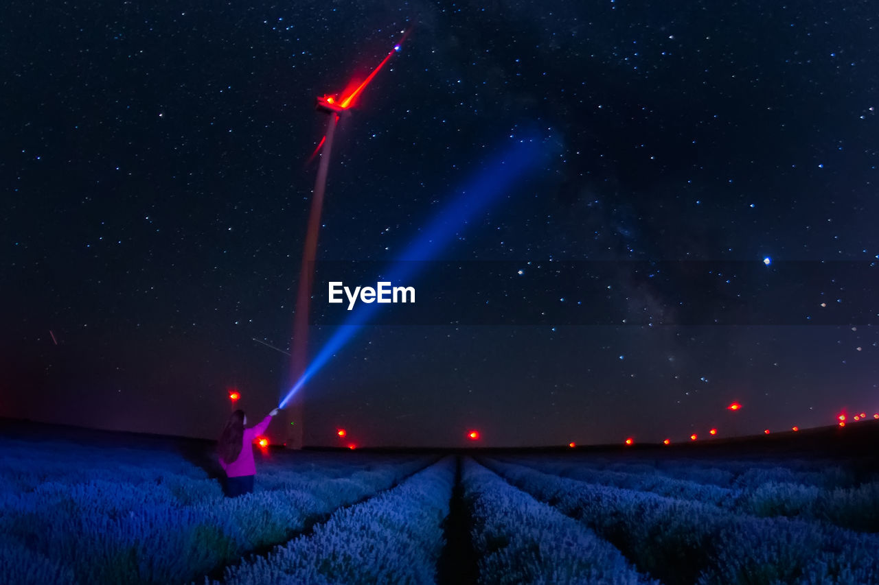 Rear view of woman holding flashlight standing on field against sky at night