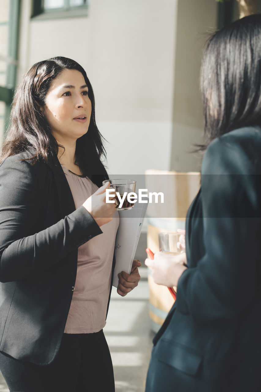 Female professionals having coffee while discussing in office corridor