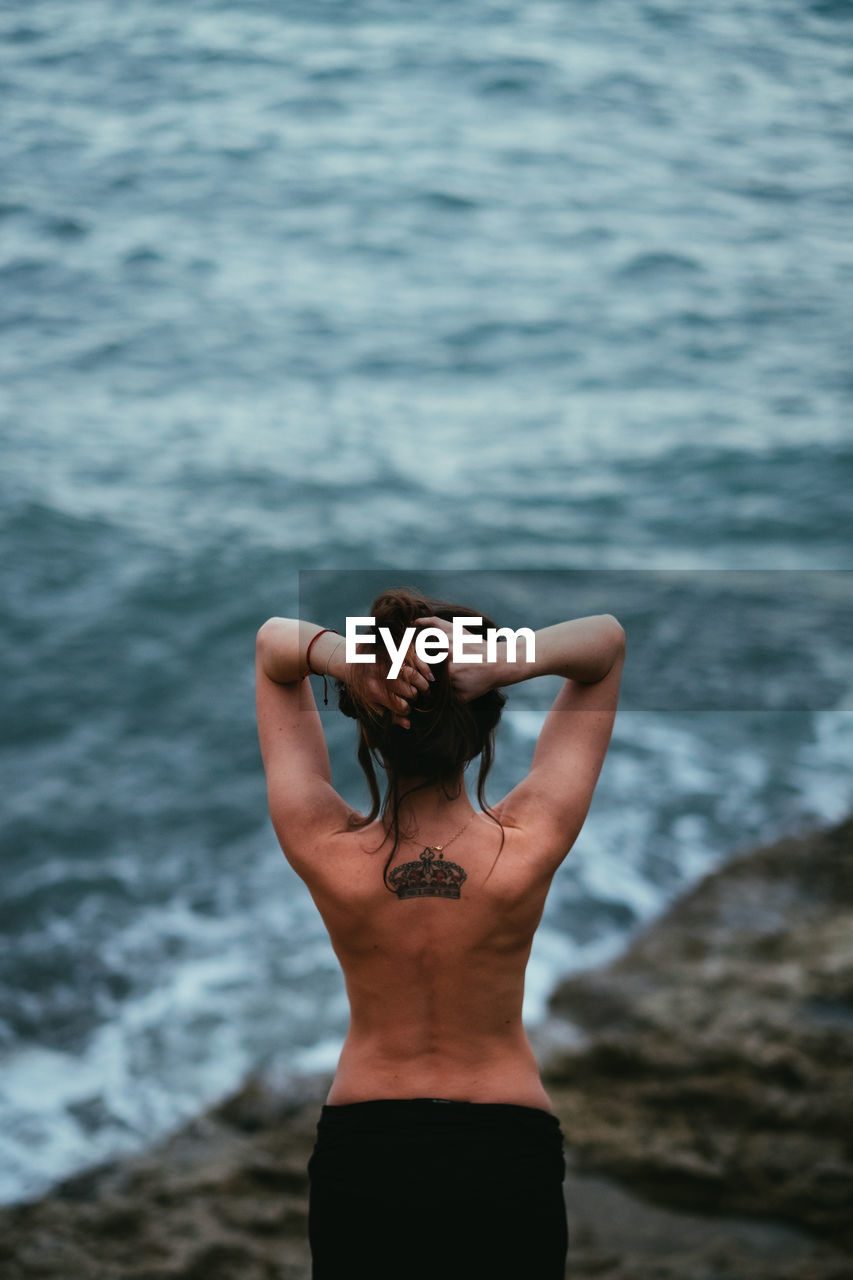 Rear view of shirtless young woman standing on rock at beach