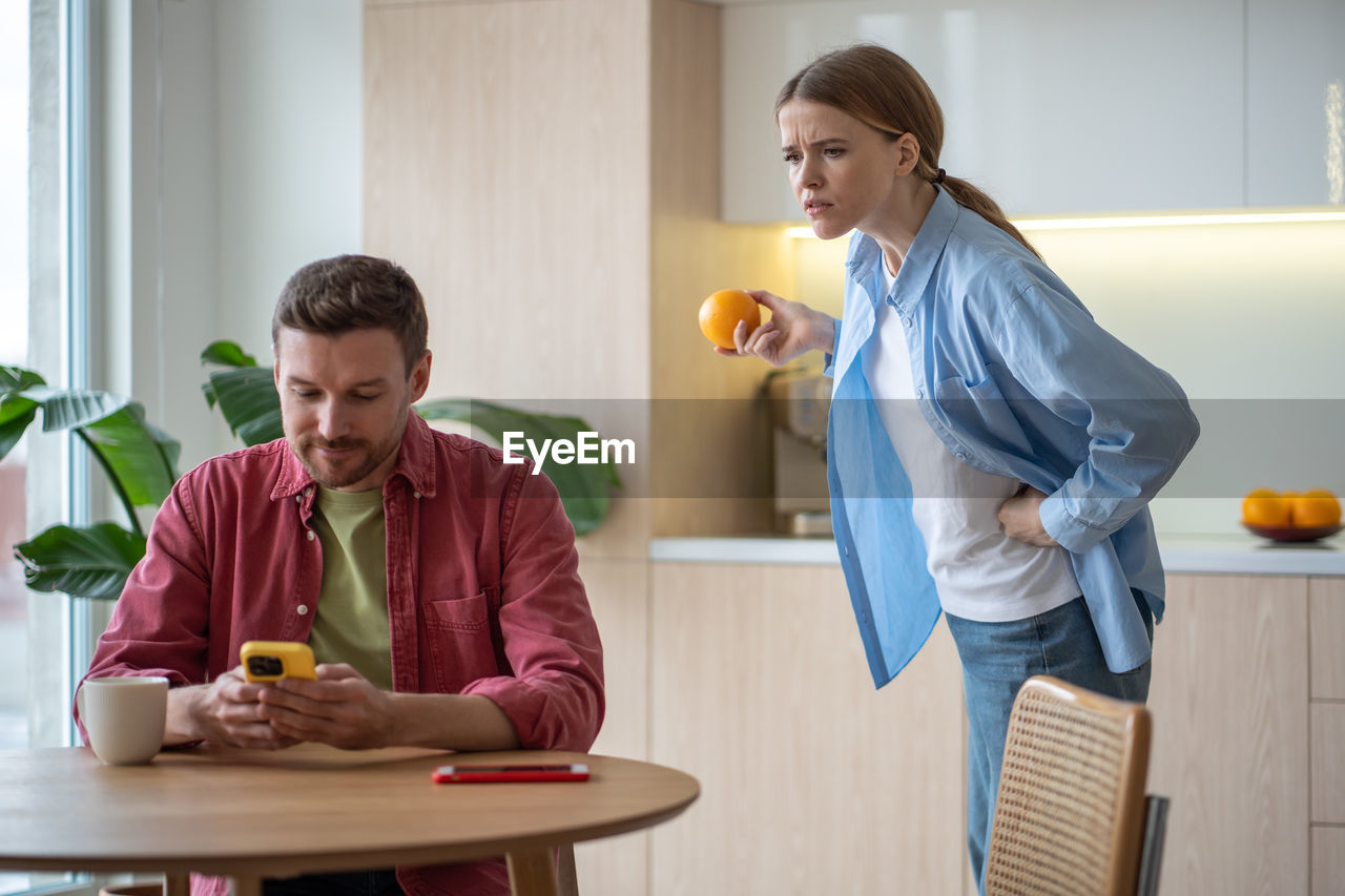 Frowning woman peeks into smartphone of grinning man, suspecting of communicating with another woman