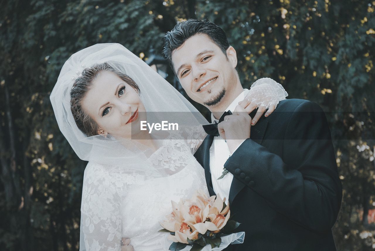 Portrait of happy young bride and groom standing against trees