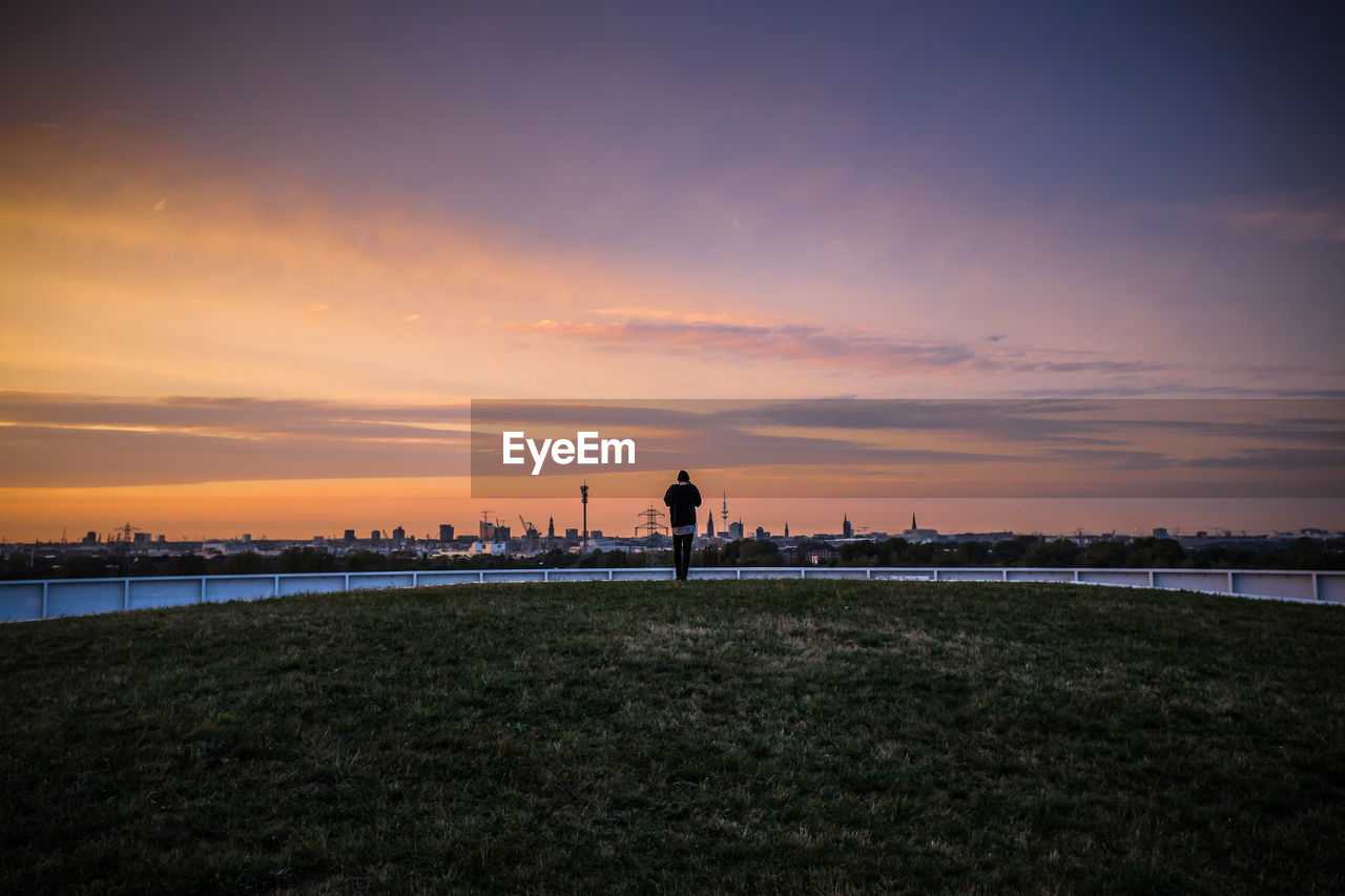 Rear view of person standing on grassy field against sky during sunset