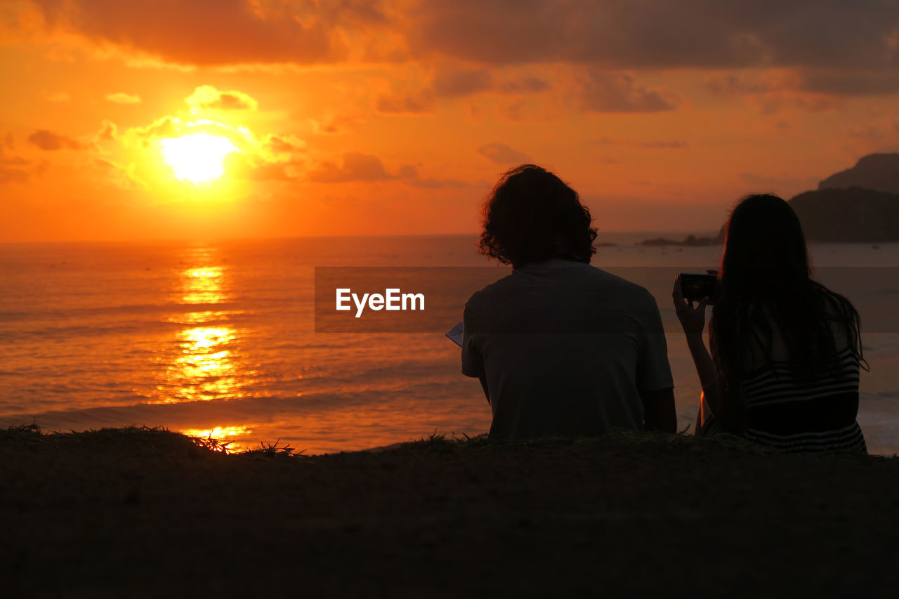 REAR VIEW OF MEN SITTING ON BEACH AGAINST SUNSET SKY