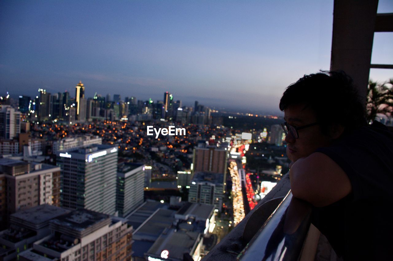 Side view of young man looking at illuminated cityscape through window at home