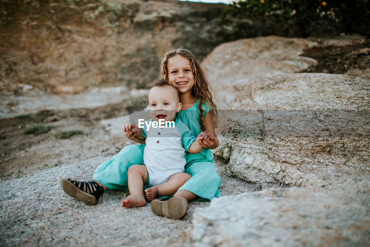 Happy young siblings sitting together on rock smiling