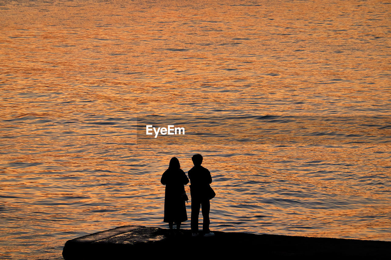 SILHOUETTE MEN STANDING ON SHORE AGAINST SEA DURING SUNSET
