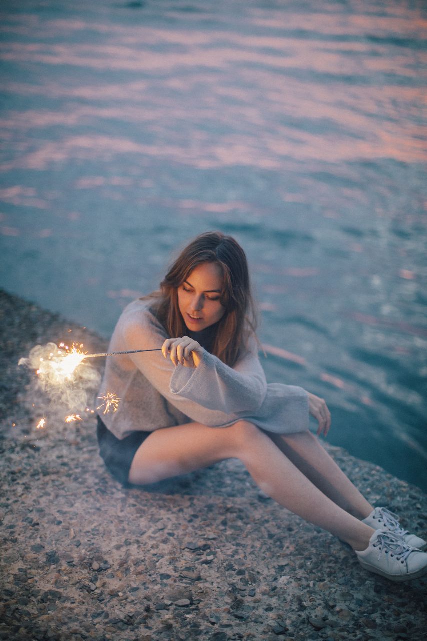 Woman with lit sparkler sitting against lake