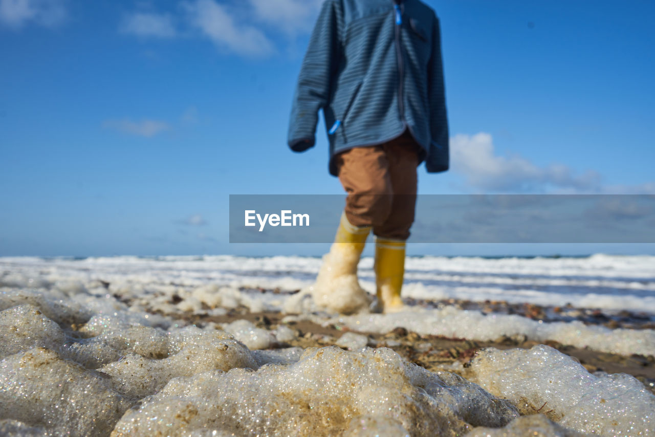 LOW SECTION OF MAN STANDING ON BEACH AGAINST SKY