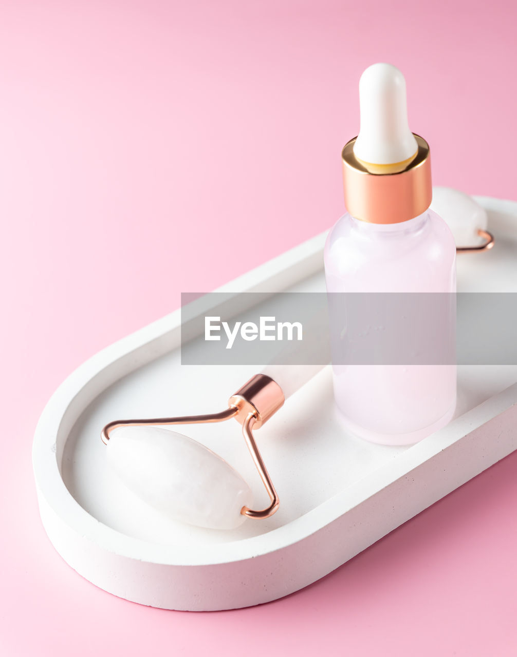 Cosmetic serum in glass bottle and face roller on white concrete tray on pink background. skincare