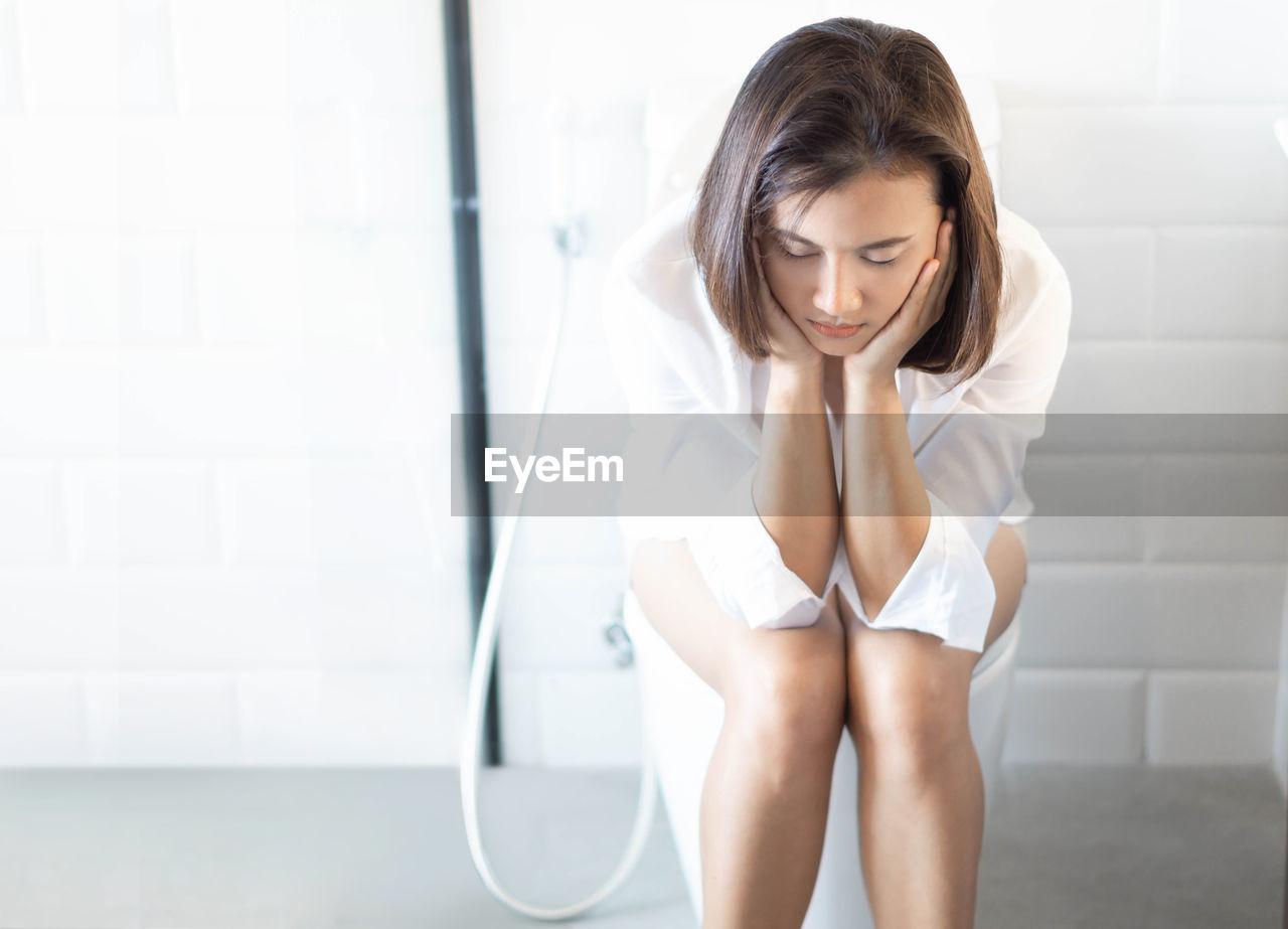 Sad woman sitting on toilet bowl in bathroom at home