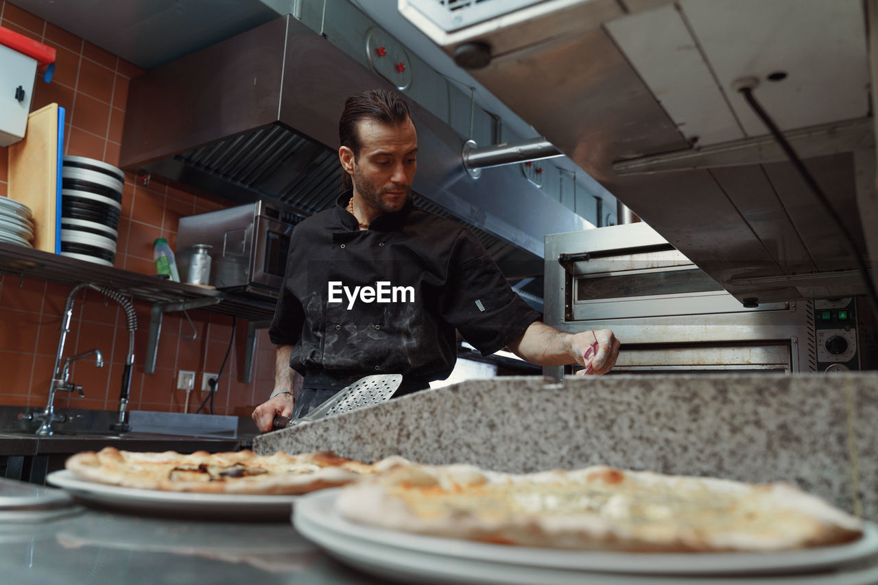 Portrait of young male chef standing in kitchen and preparing pizza