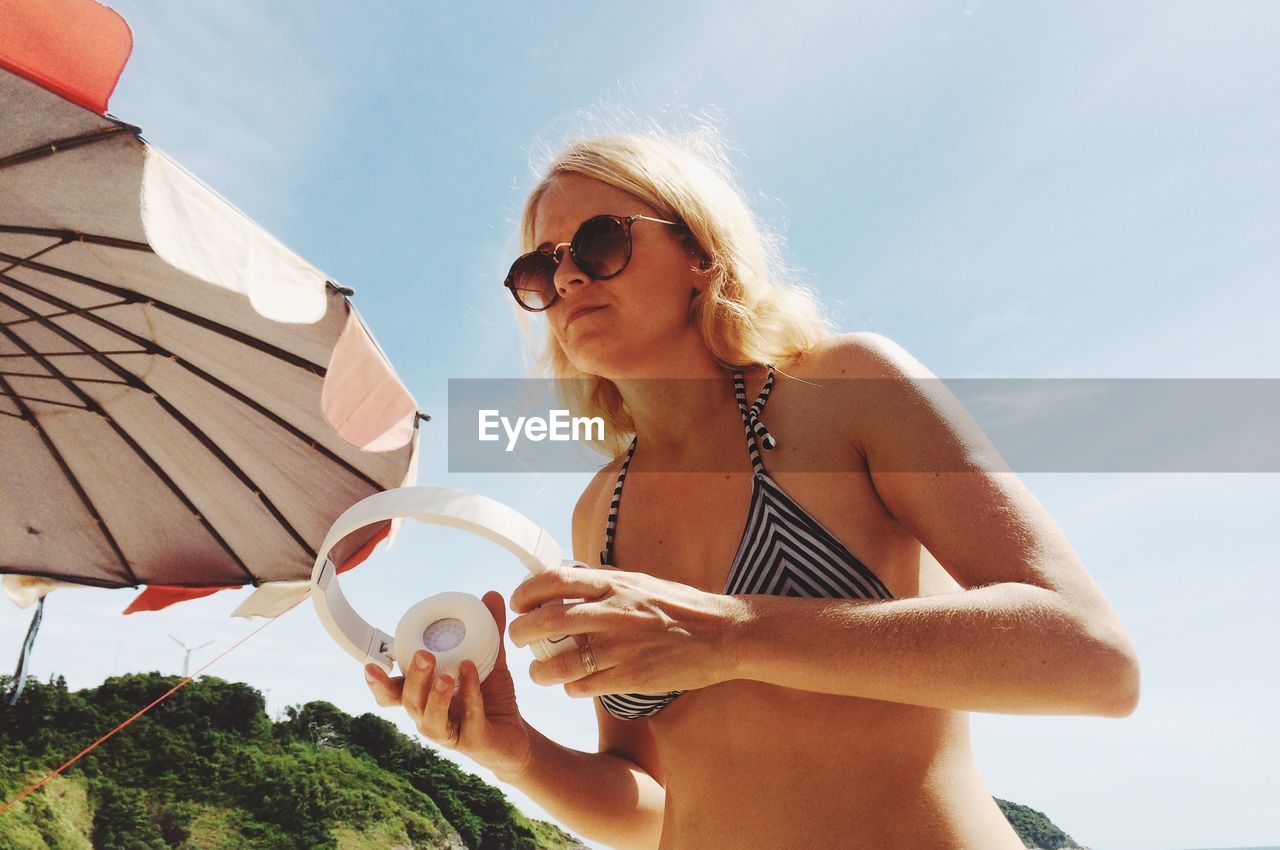 Low angle view of woman wearing sunglasses and bikini top holding headphones against sky during sunny day