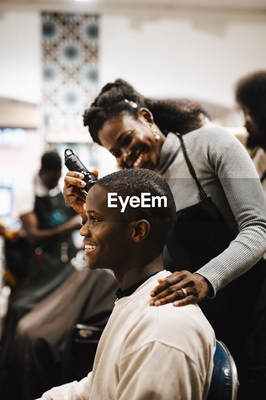 Smiling female barber cutting hair of male customer with electric razor at salon