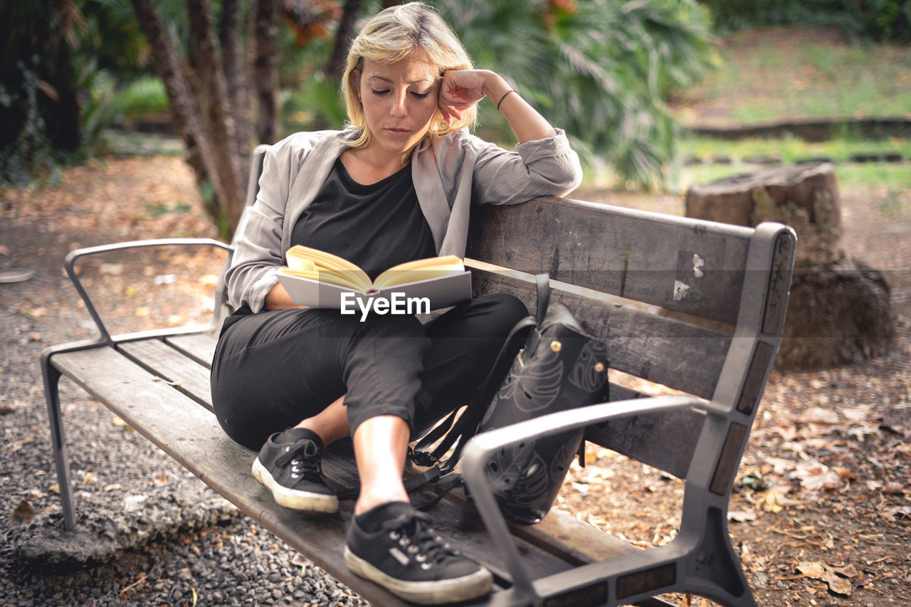 Full length of woman reading book sitting on bench at park