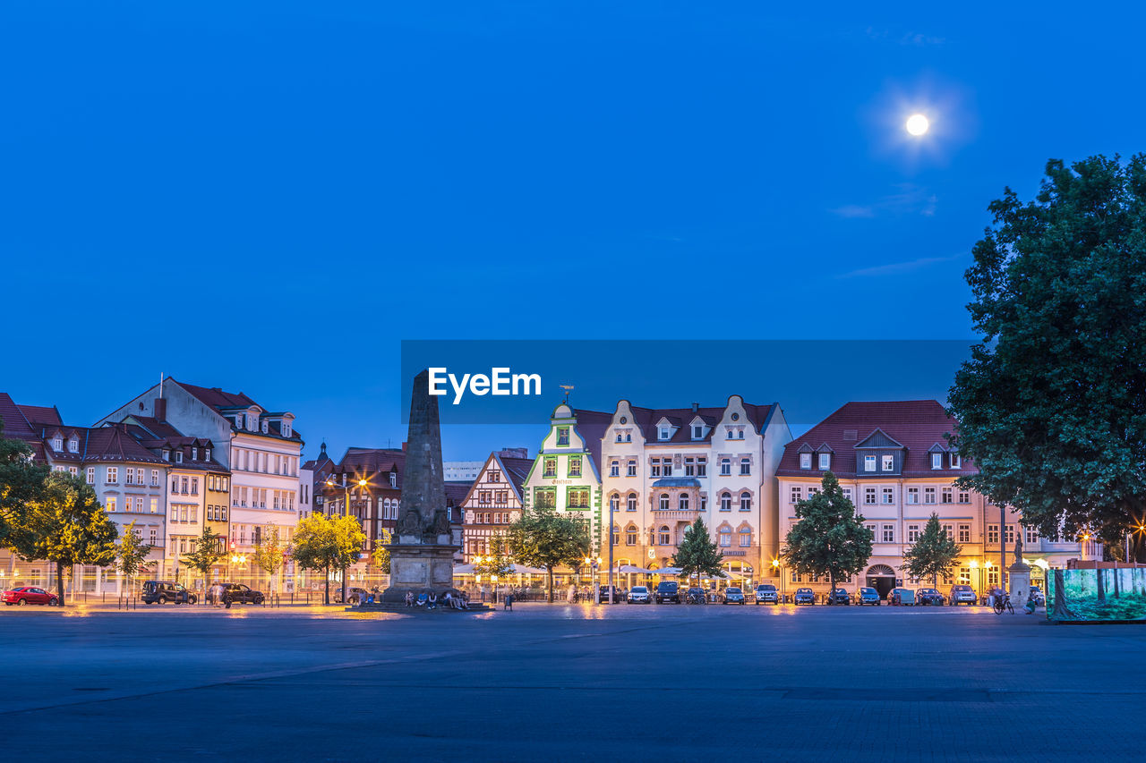 Illuminated buildings by street against blue sky at night