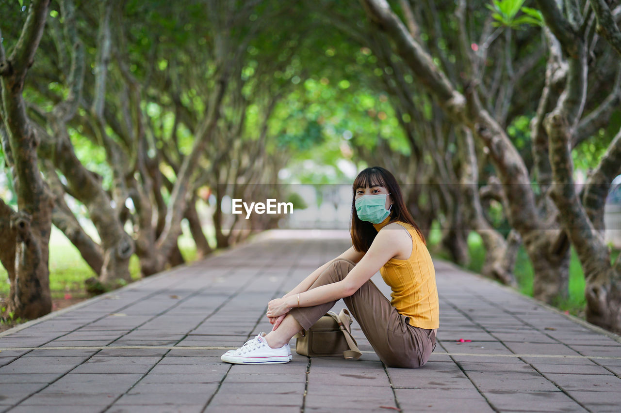 Portrait of young woman wearing mask and sitting by tree