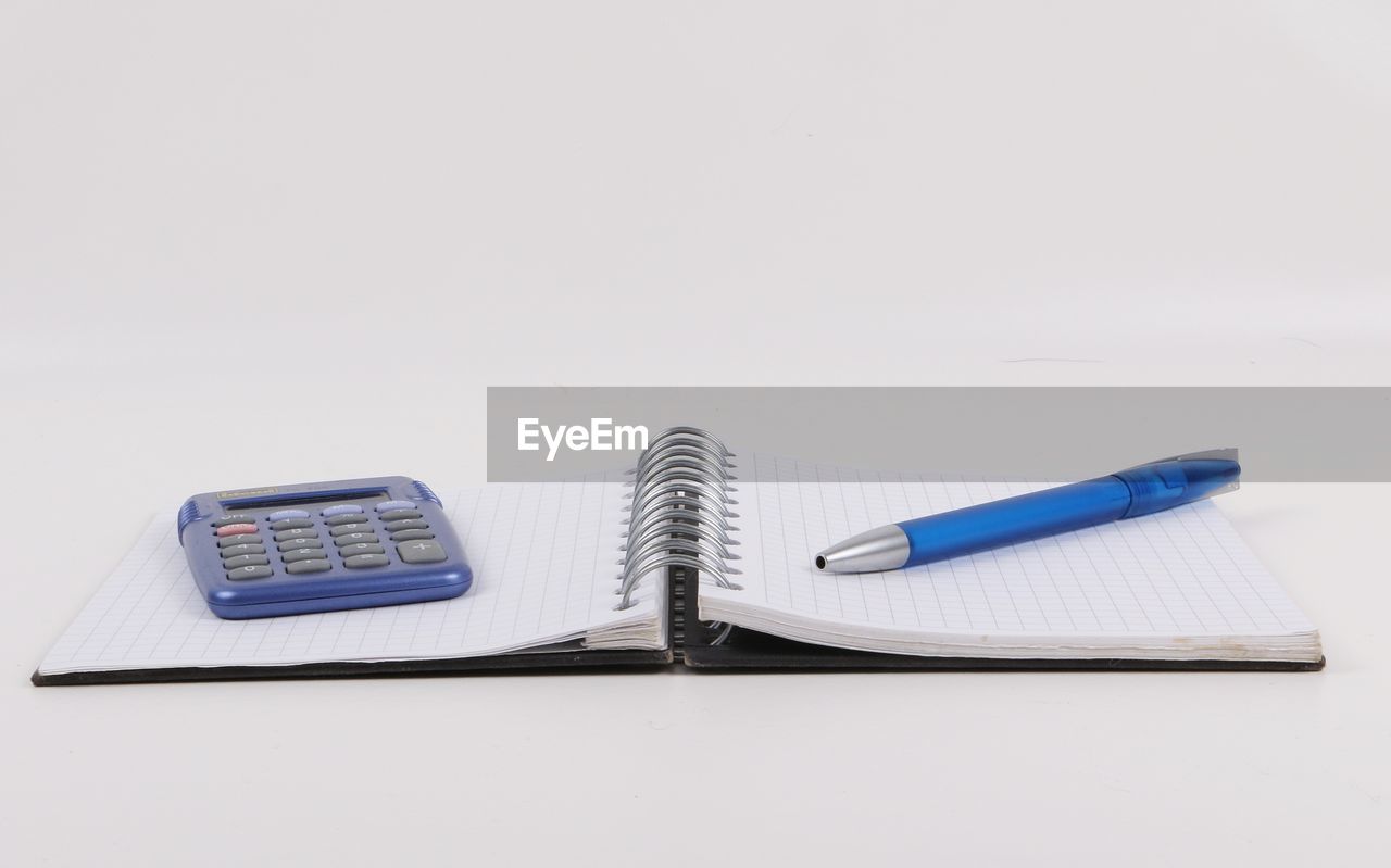 Close-up of spiral notebook with pen and calculator on white background