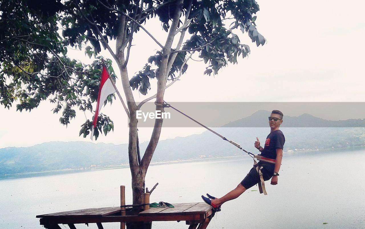 Young man swinging from tree by river against clear sky