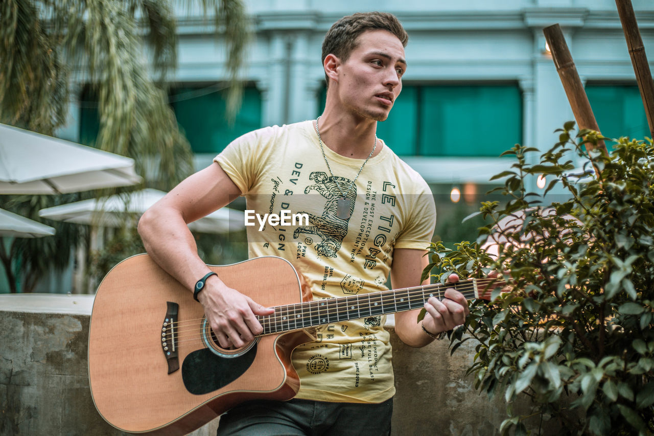 Thoughtful young man playing guitar while standing against building