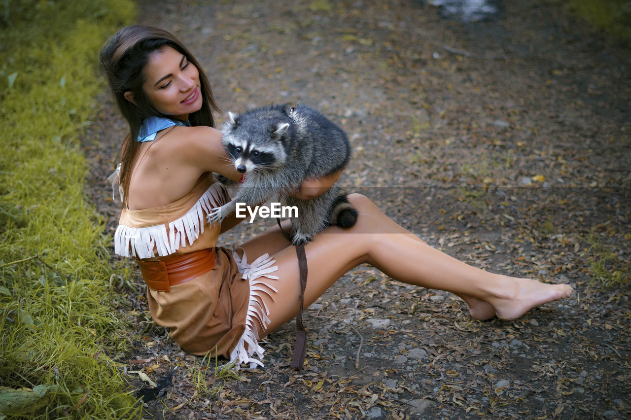 Young woman in traditional clothing playing with raccoon in forest