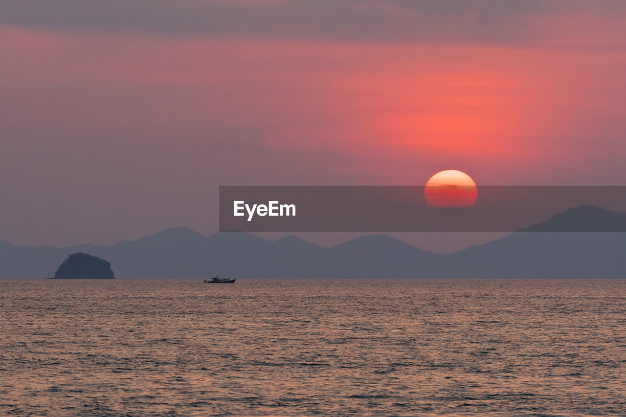 Red circle of sun in a haze sets behind the silhouette of the mountains and on sea horizon