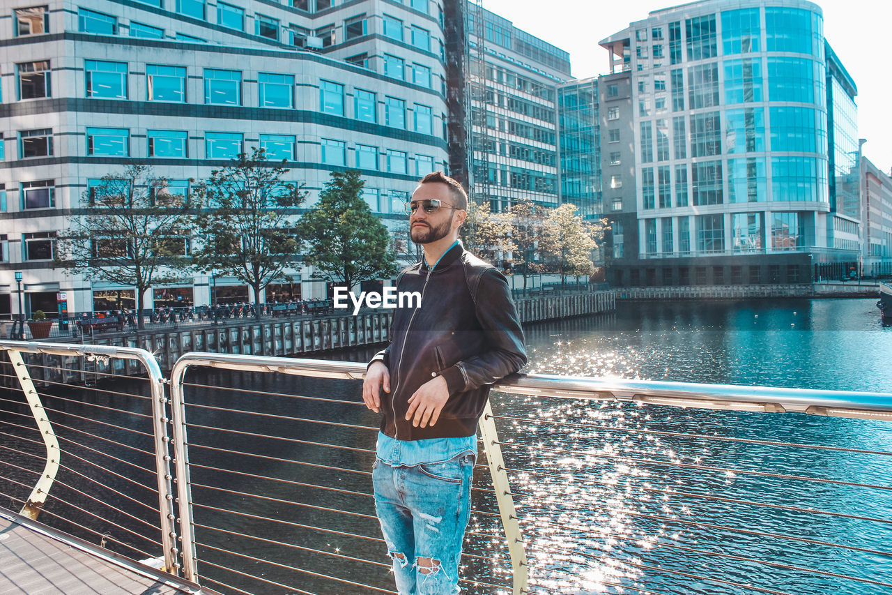 Young man wearing sunglasses standing on bridge over river in city