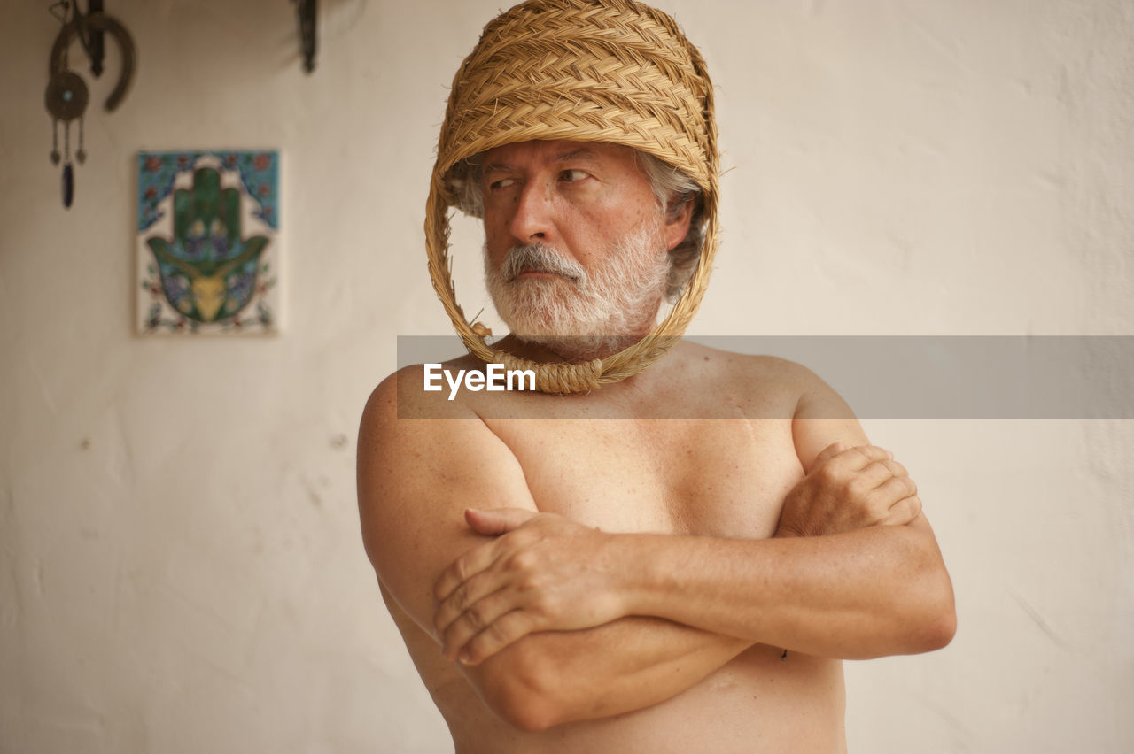 Shirtless man wearing basket while standing against wall