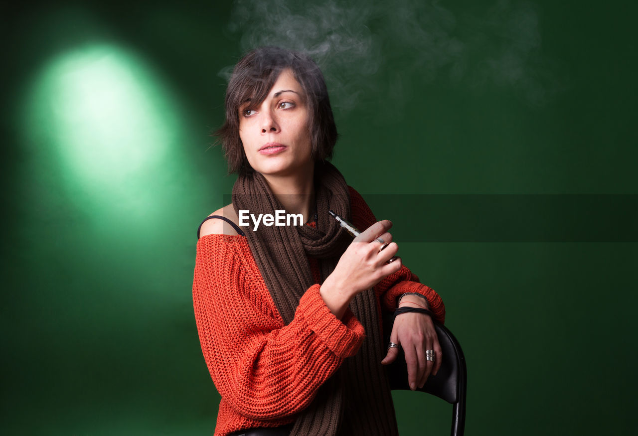 Young woman using electric cigarette while sitting against green background