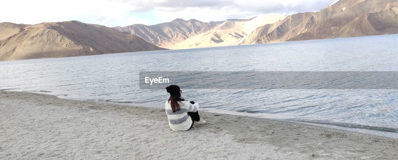 Rear view of woman sitting at lakeshore by mountains