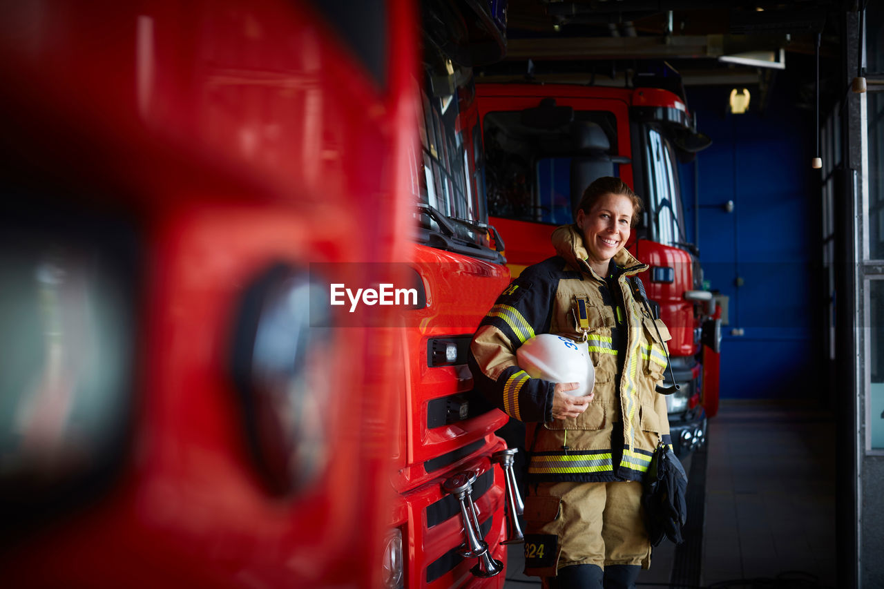 Portrait of smiling firefighter with helmet standing by fire engine at fire station