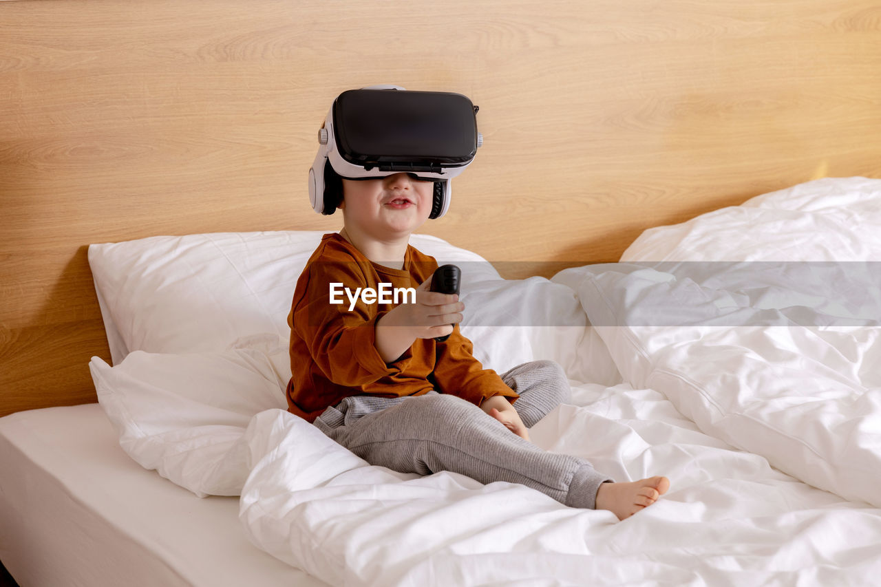 Little adorable boy sitting on bed at home with vr headset and playing interactive video game
