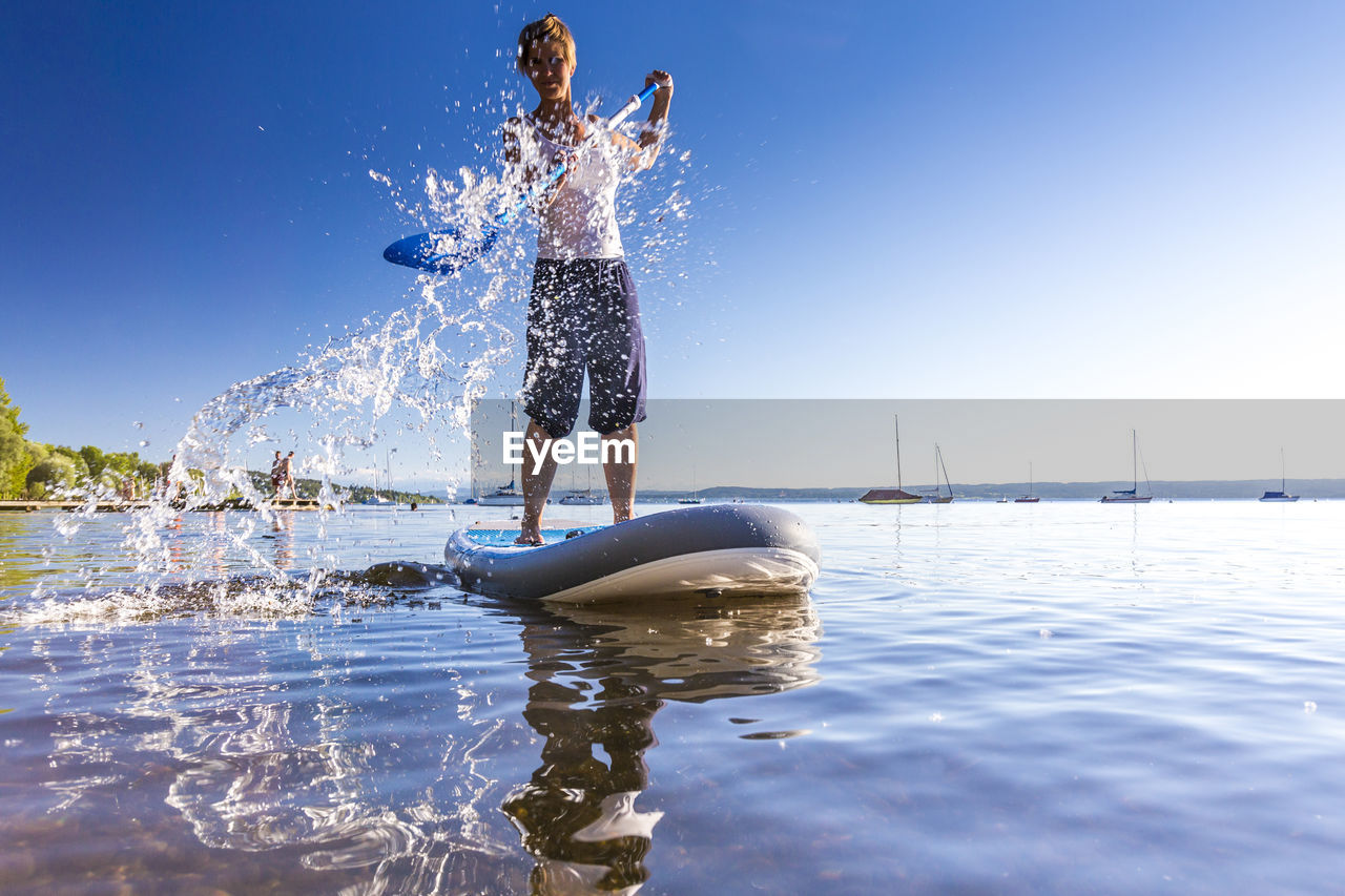 Full length of young woman paddleboarding in lake against clear blue sky