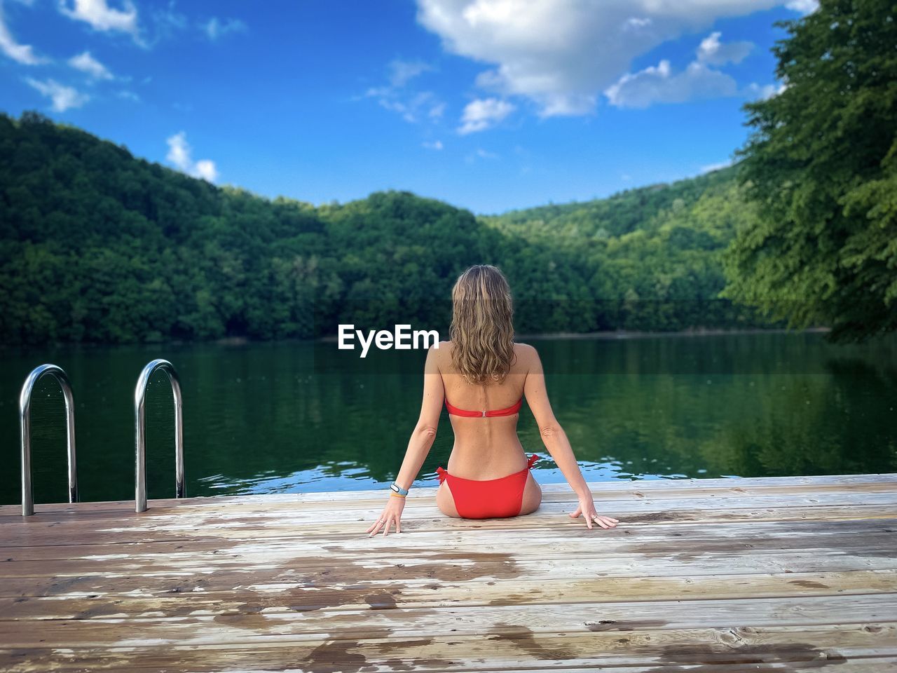 Woman in red bikini sitting down on a wooden pontoon looking at the lake surrounded by green forest