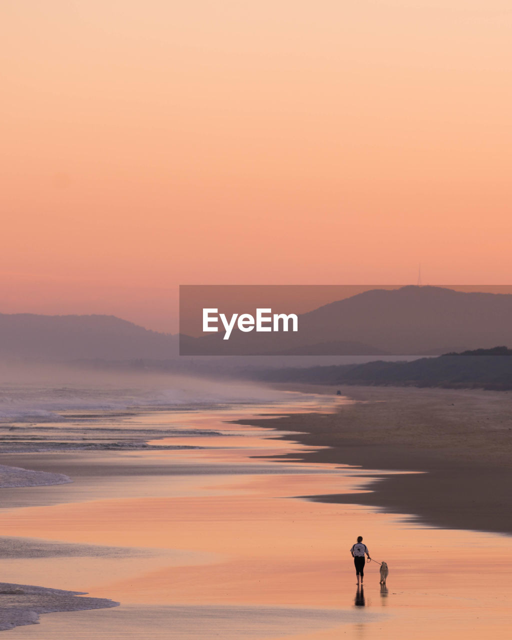 Woman standing on sea shore against orange sky