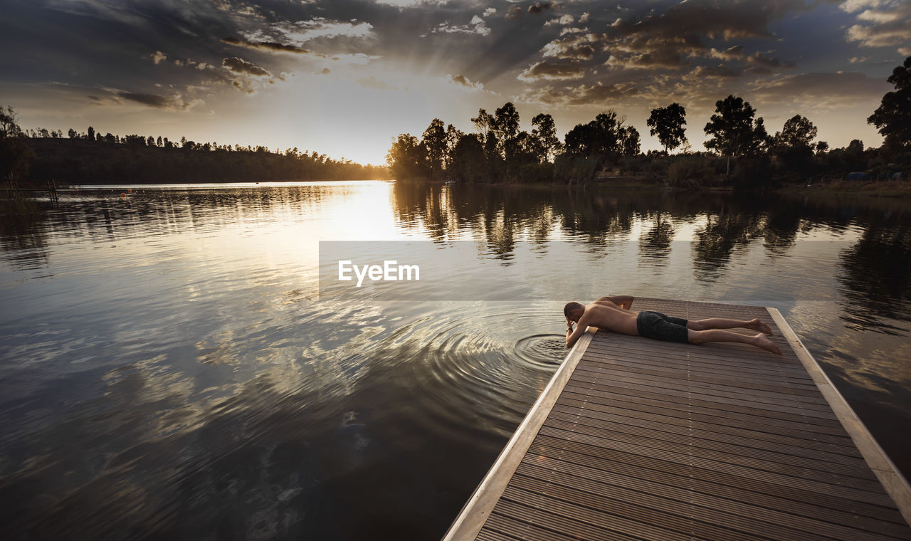 Shirtless man practicing yoga on pier against sky during sunset