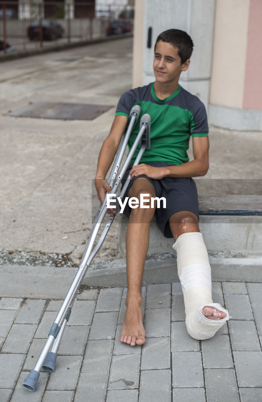 Boy with broken leg holding walking cane while sitting on footpath