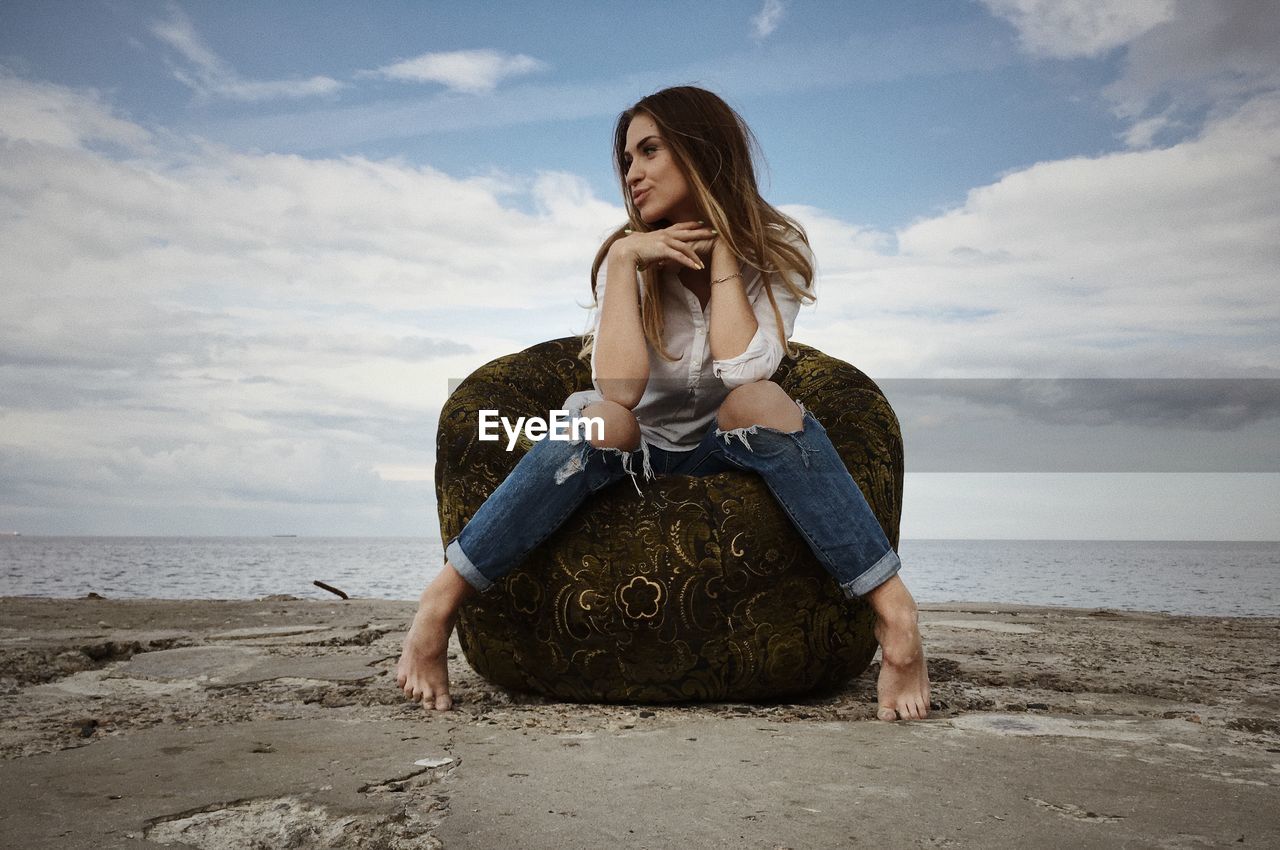 Young woman sitting on chair by sea against cloudy sky