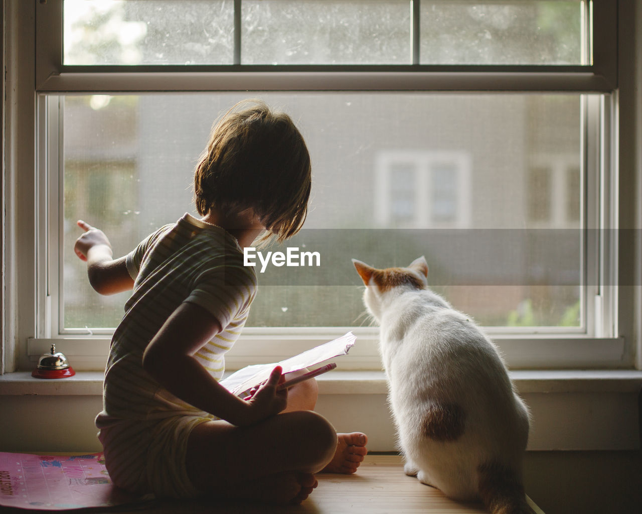 A little girl sits on a bench with a cat looking out window together