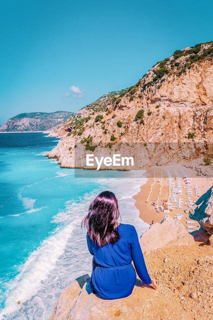 Rear view of woman sitting on cliff against beach