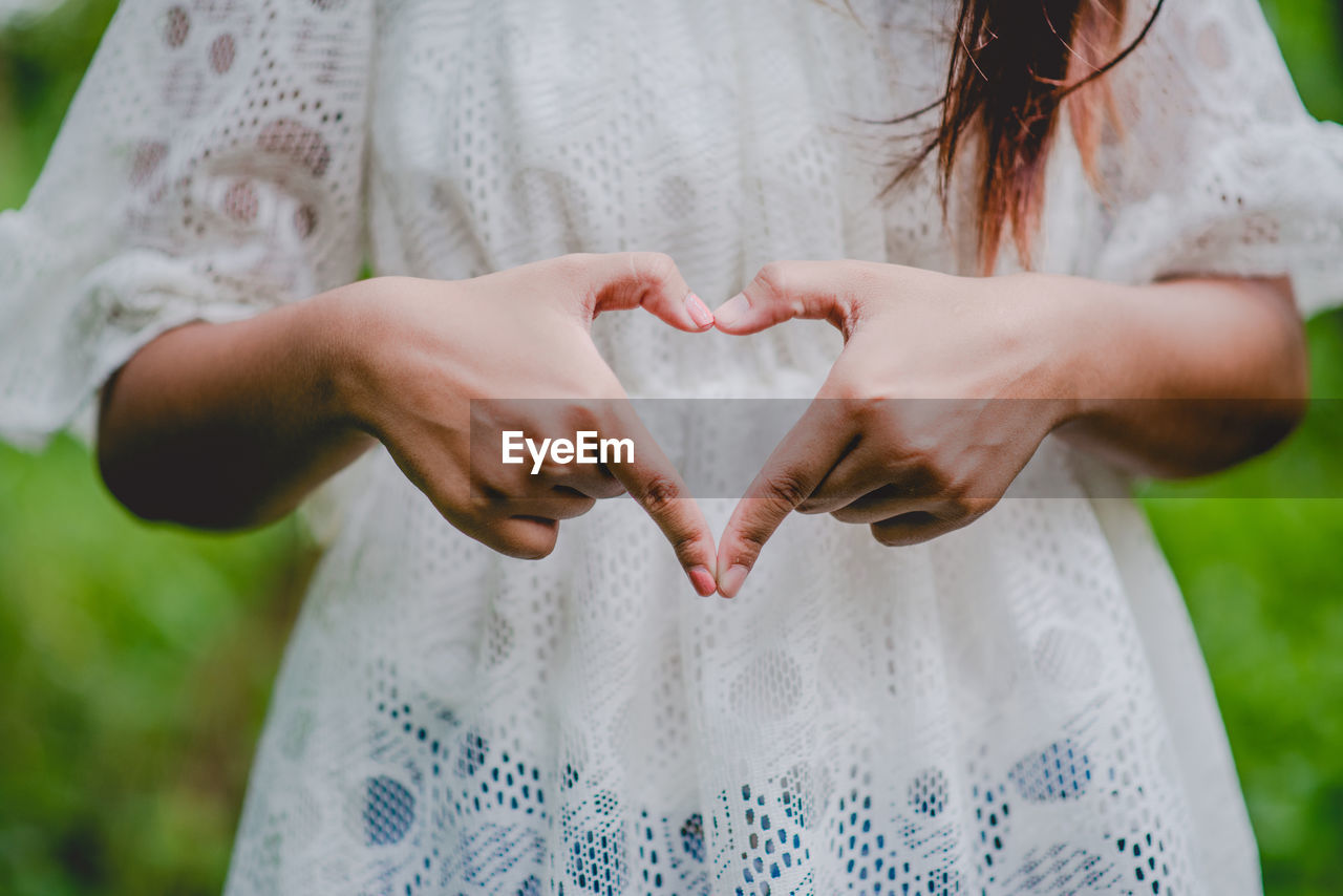 MIDSECTION OF WOMAN MAKING HEART SHAPE AGAINST WHITE WALL