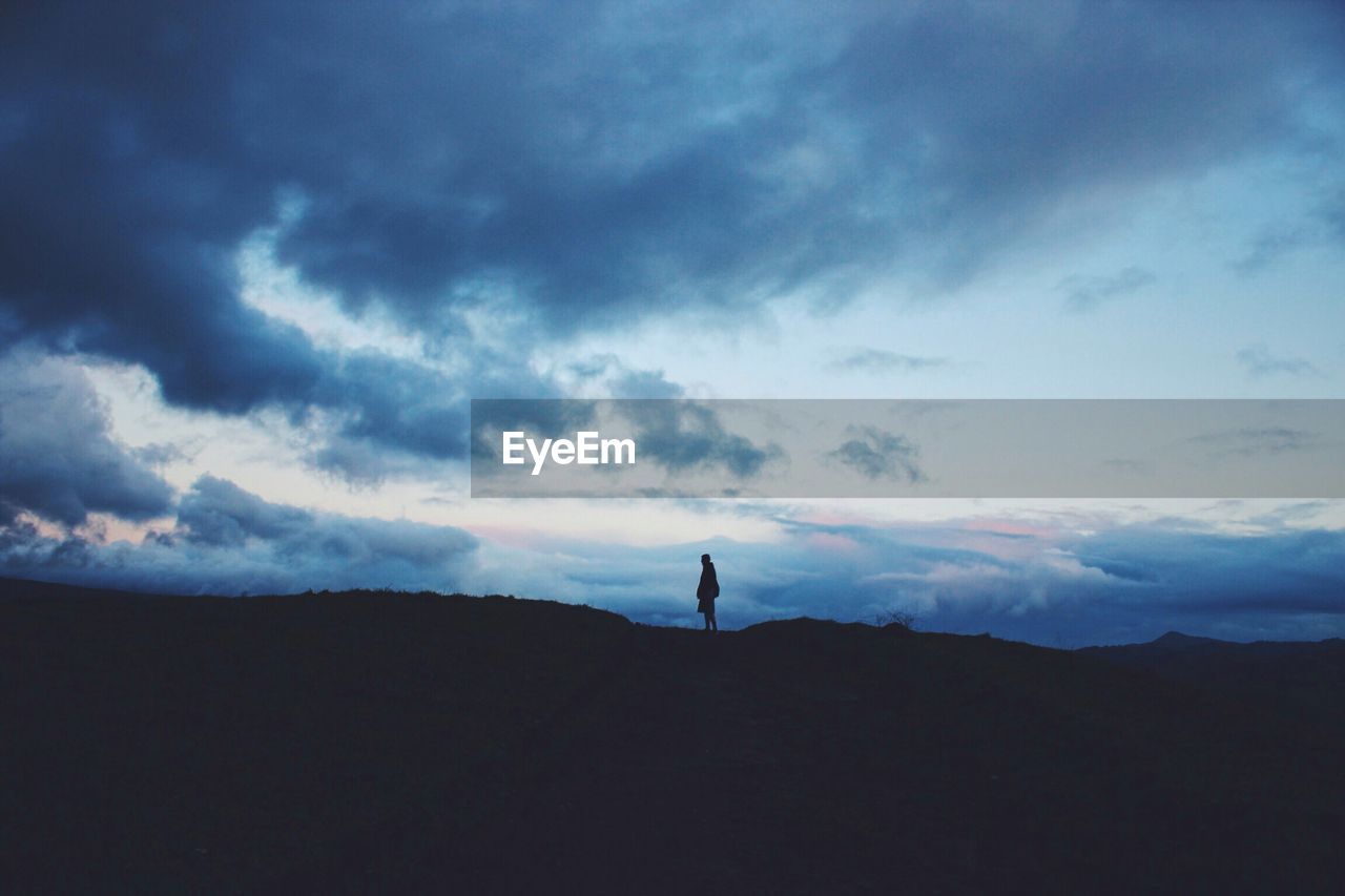 Silhouette person standing on hill against cloudy sky at dusk