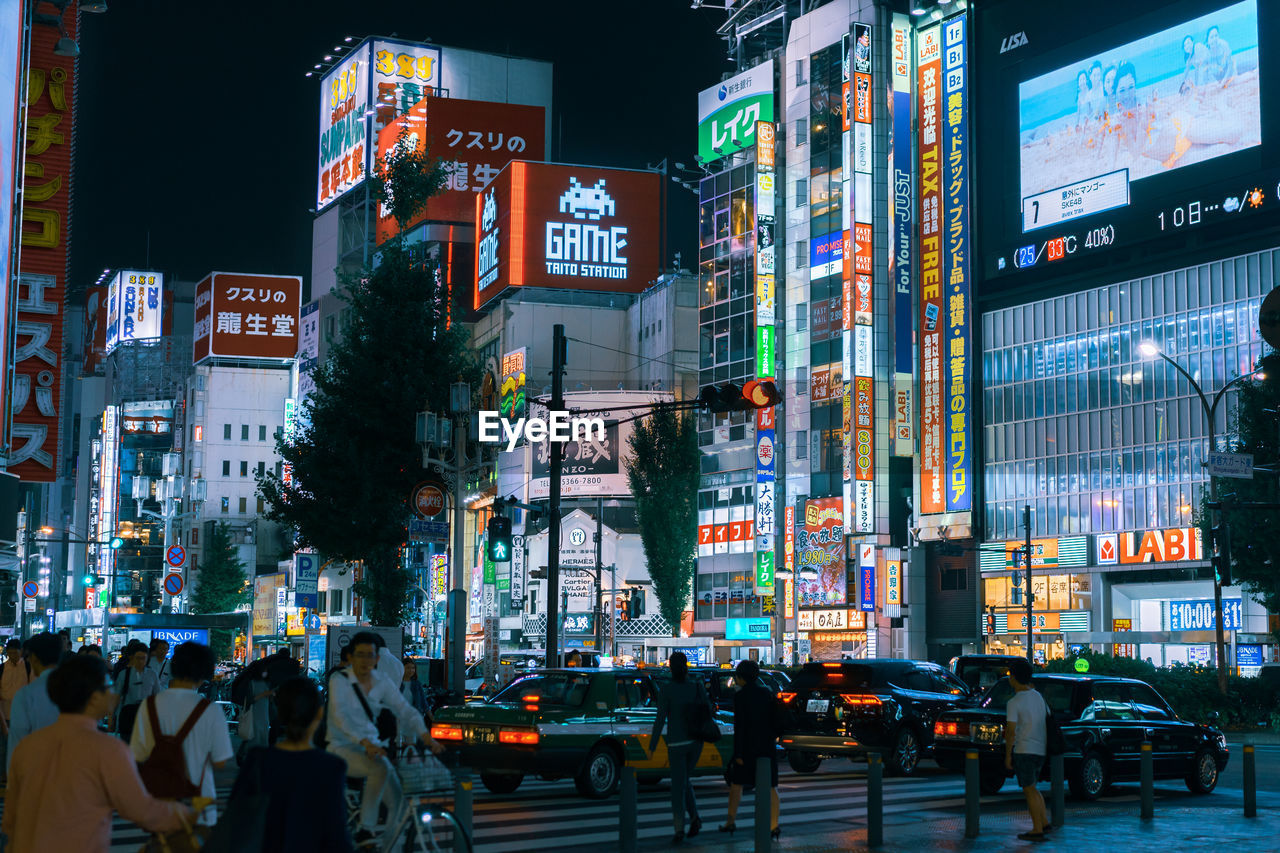 PEOPLE ON ILLUMINATED STREET AMIDST BUILDINGS IN CITY