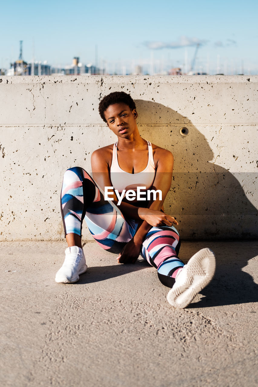 African american female athlete in flowered sport clothes and white sneakers looking at camera while sitting alone on concrete wall in sunbeams and resting after training in city