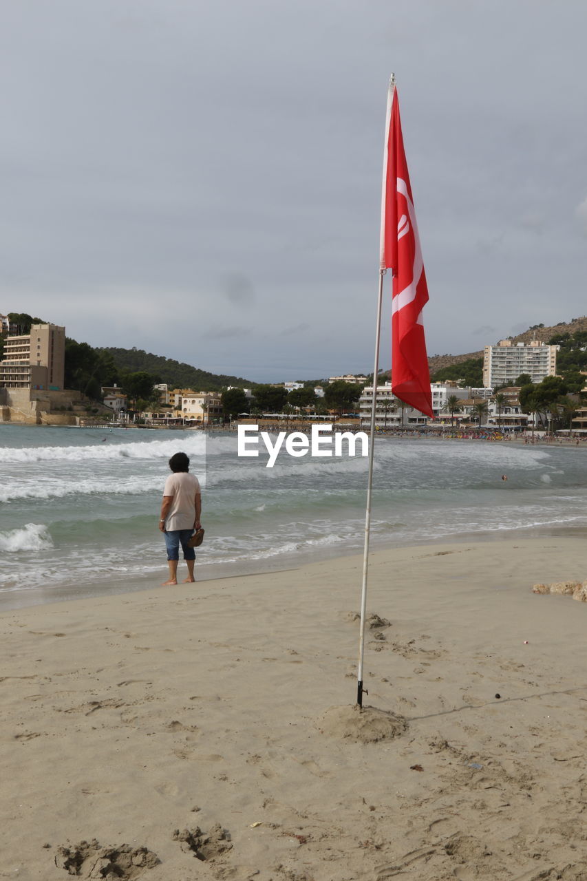 REAR VIEW OF MAN STANDING AT BEACH AGAINST SKY