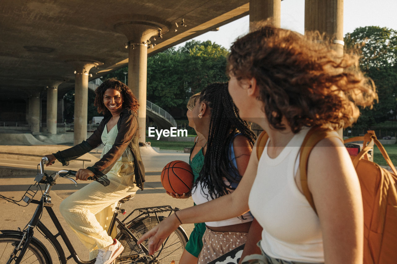 Teenage girls looking at smiling female friend riding bicycle under bridge