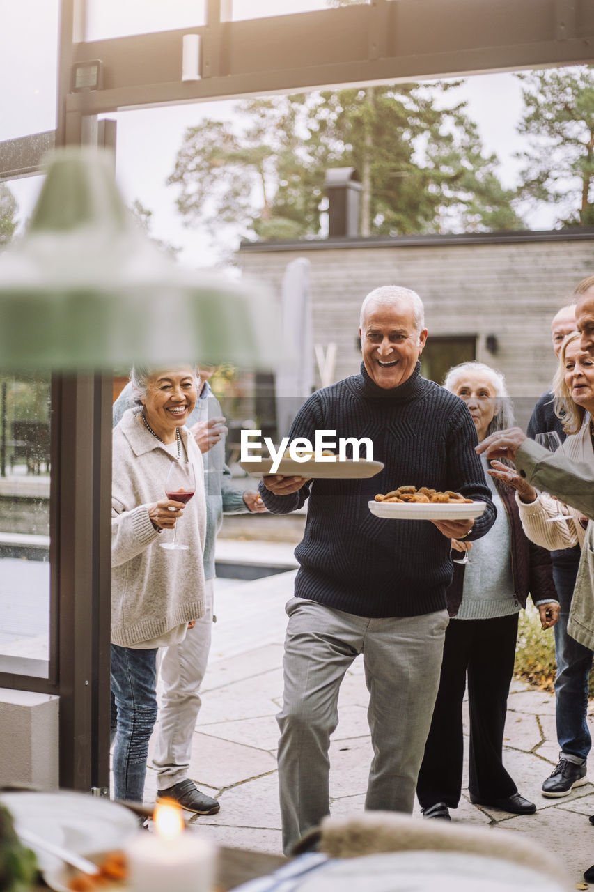 Happy mature man carrying food plates walking with friends during dinner party