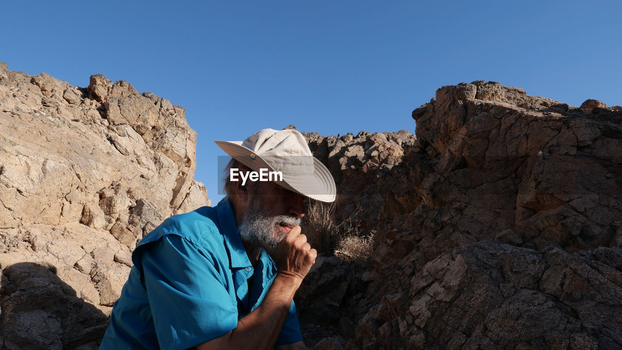 Side view of senior man sitting on rock against clear blue sky