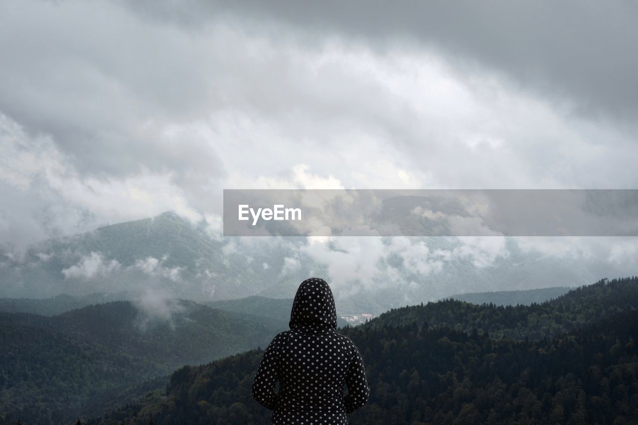 Rear view of mid adult woman looking at mountain range while standing against cloudy sky