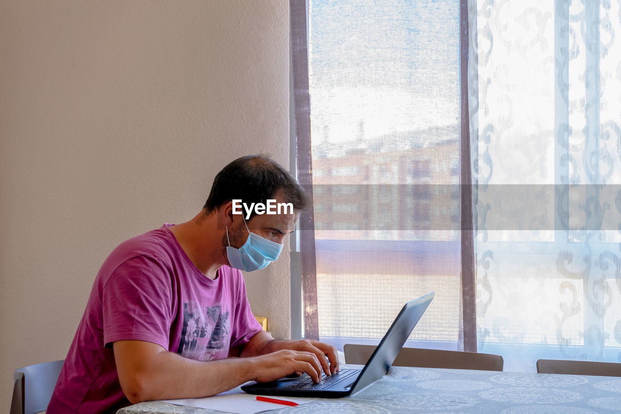 Side view of man wearing flu mask using laptop at home